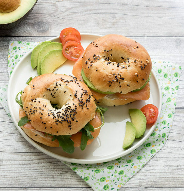 Bagel con salmone, avocado e pomodoro su piatto bianco con tovagliolo fiorato.