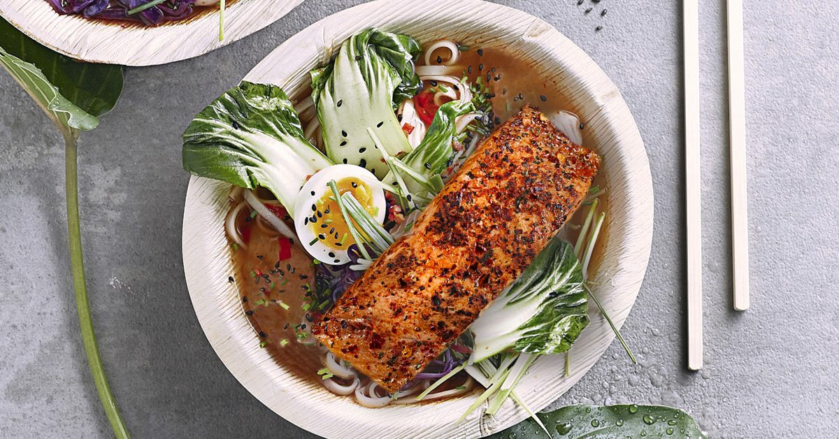 Salmone speziato su ramen con verdure e uovo, presentato su un piatto biodegradabile.
