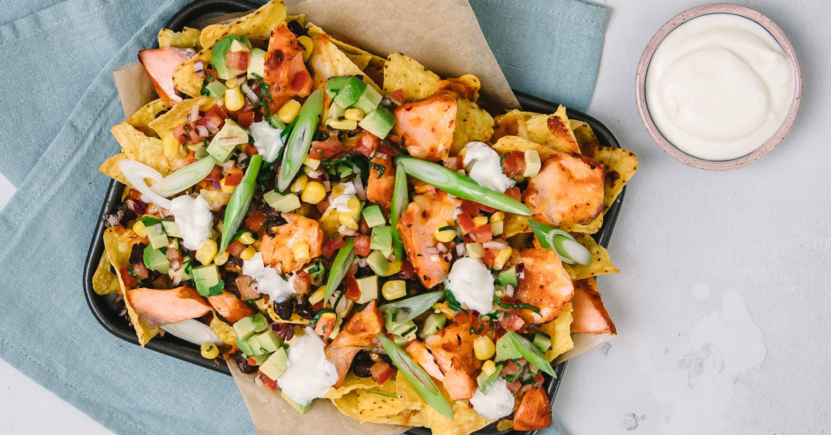 Nachos con salmone, gamberi, mais, avocado e cipollotti su una teglia con ciotola di panna acida.