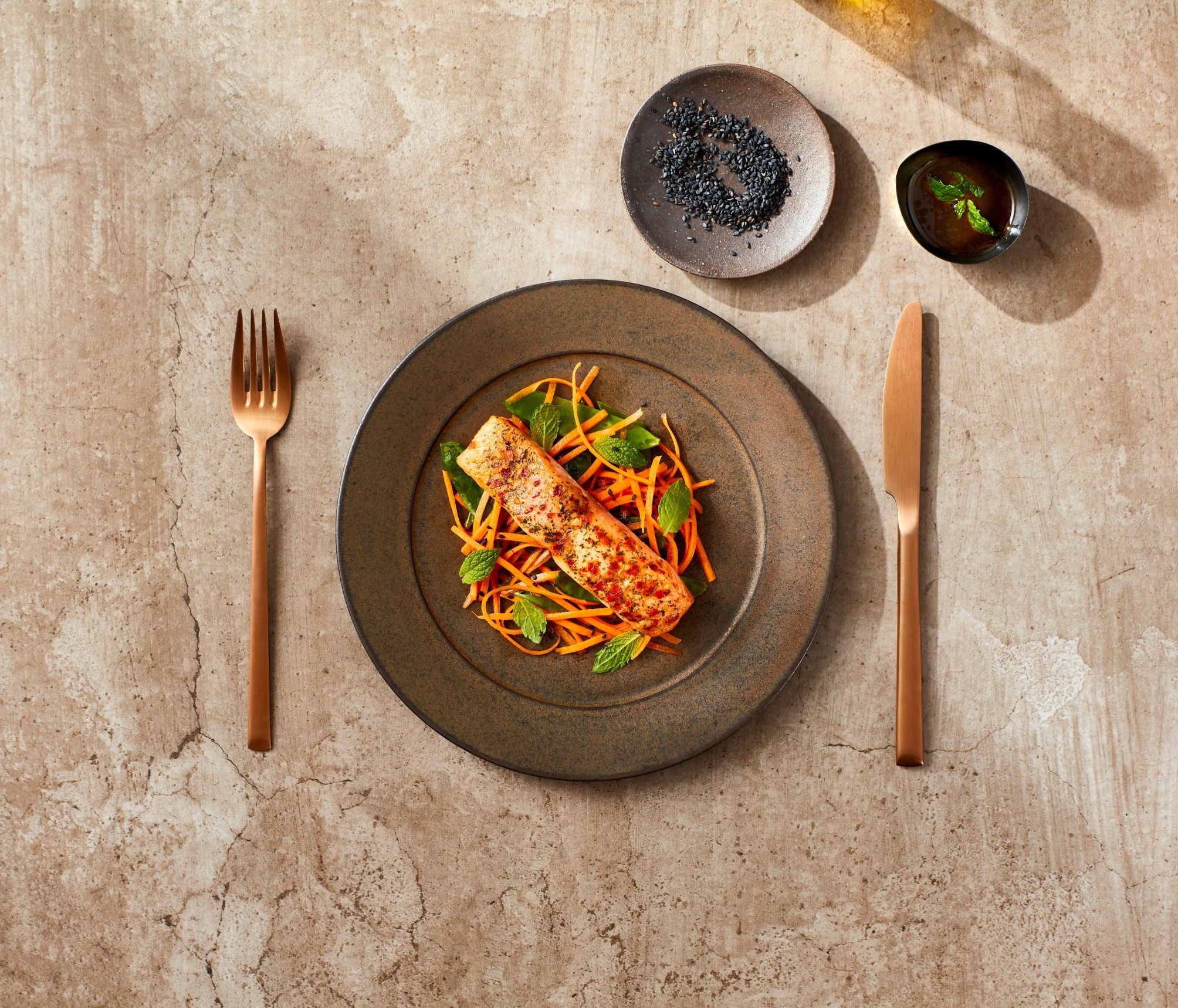 Salmone grigliato con julienne di carote e foglie di menta su un piatto di ceramica scura.