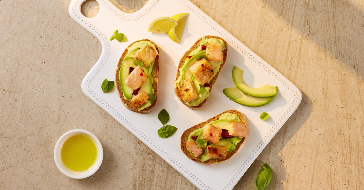 Bruschette integrali con salmone, avocado e limone su vassoio bianco.