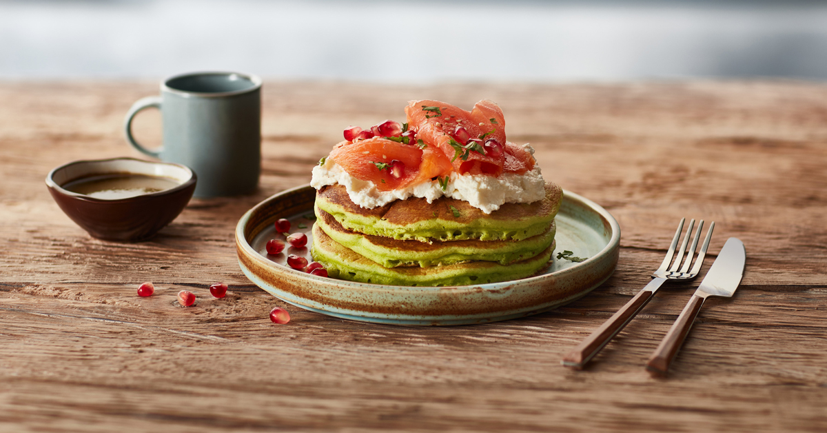 Pancakes verdi con salmone, formaggio e melograno, serviti con una tazza di caffè.