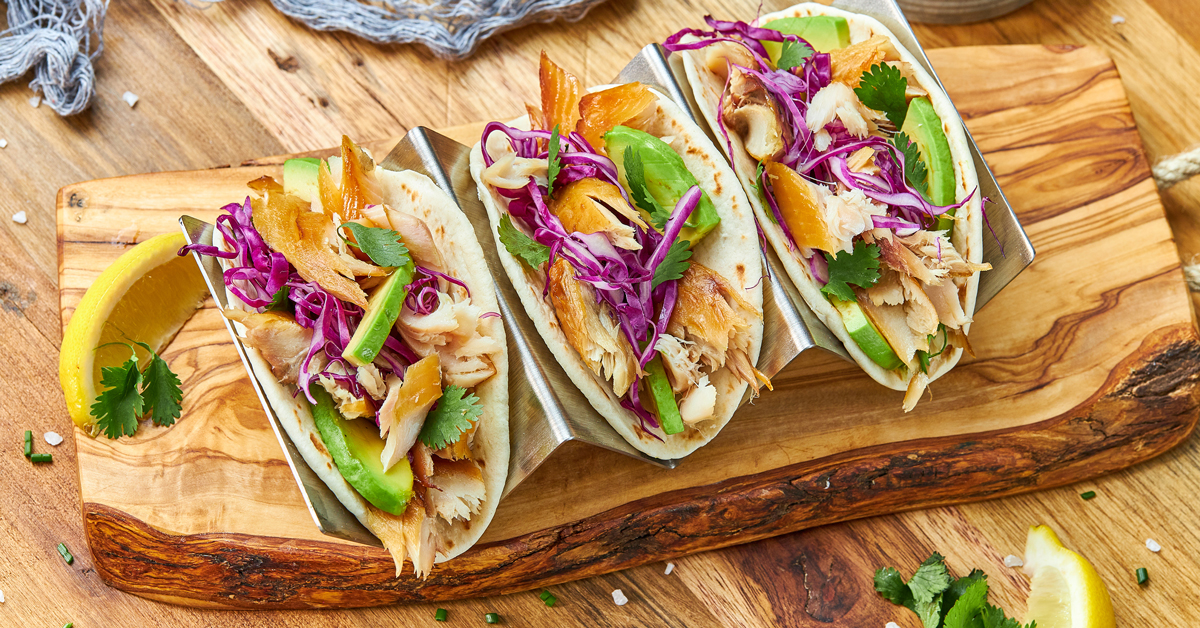 Tre tacos con pesce affumicato, cavolo rosso e avocado su un tagliere di legno.