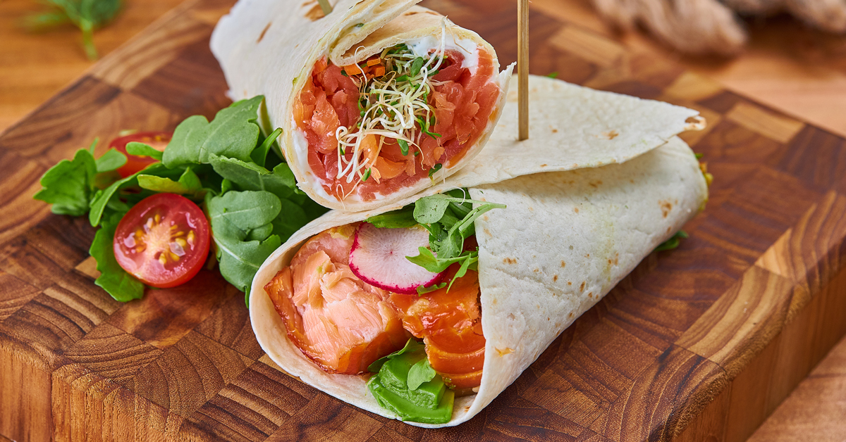 Wraps di salmone affumicato con verdure e pomodorini su tagliere di legno.