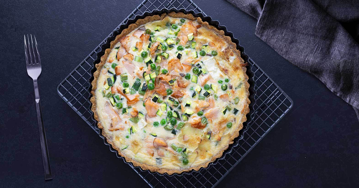 Torta salata con salmone, zucchine e piselli su griglia nera.