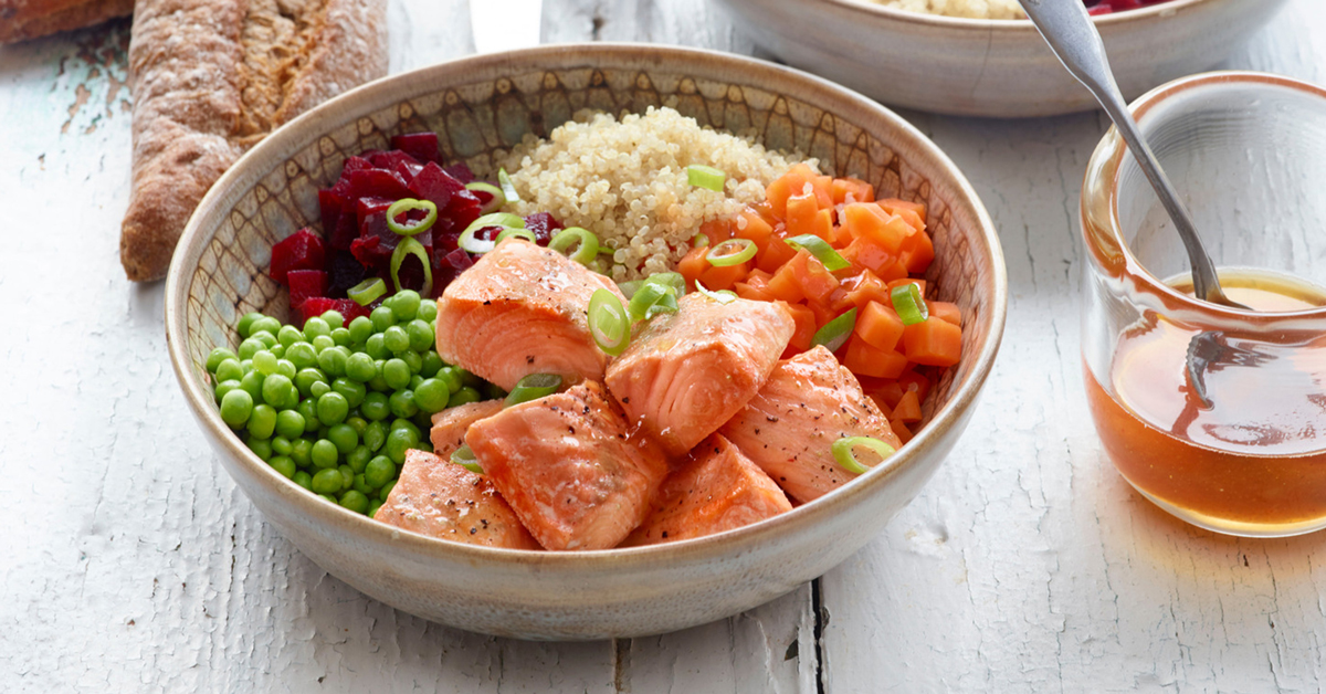 Salmone con quinoa, piselli, barbabietola e carote in una ciotola, accanto a una caraffa di miele.