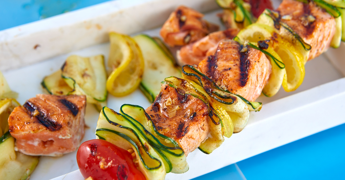 Spiedini di salmone grigliato con zucchine e limone su piatto bianco.