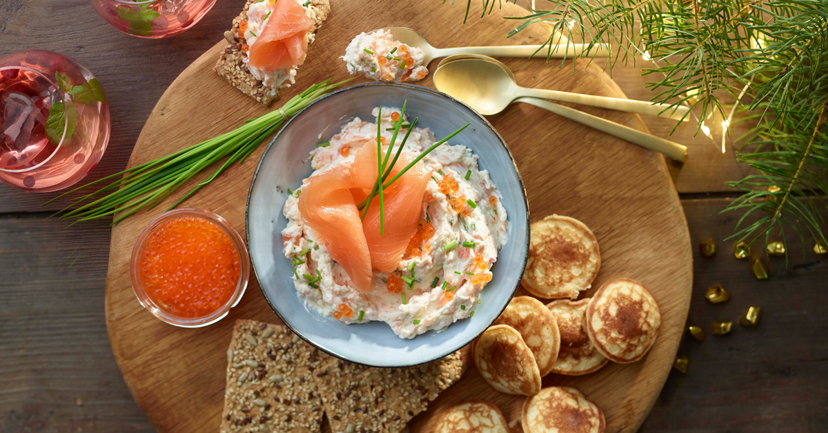 Insalata di salmone, uova di pesce e mini pancake su tagliere di legno.