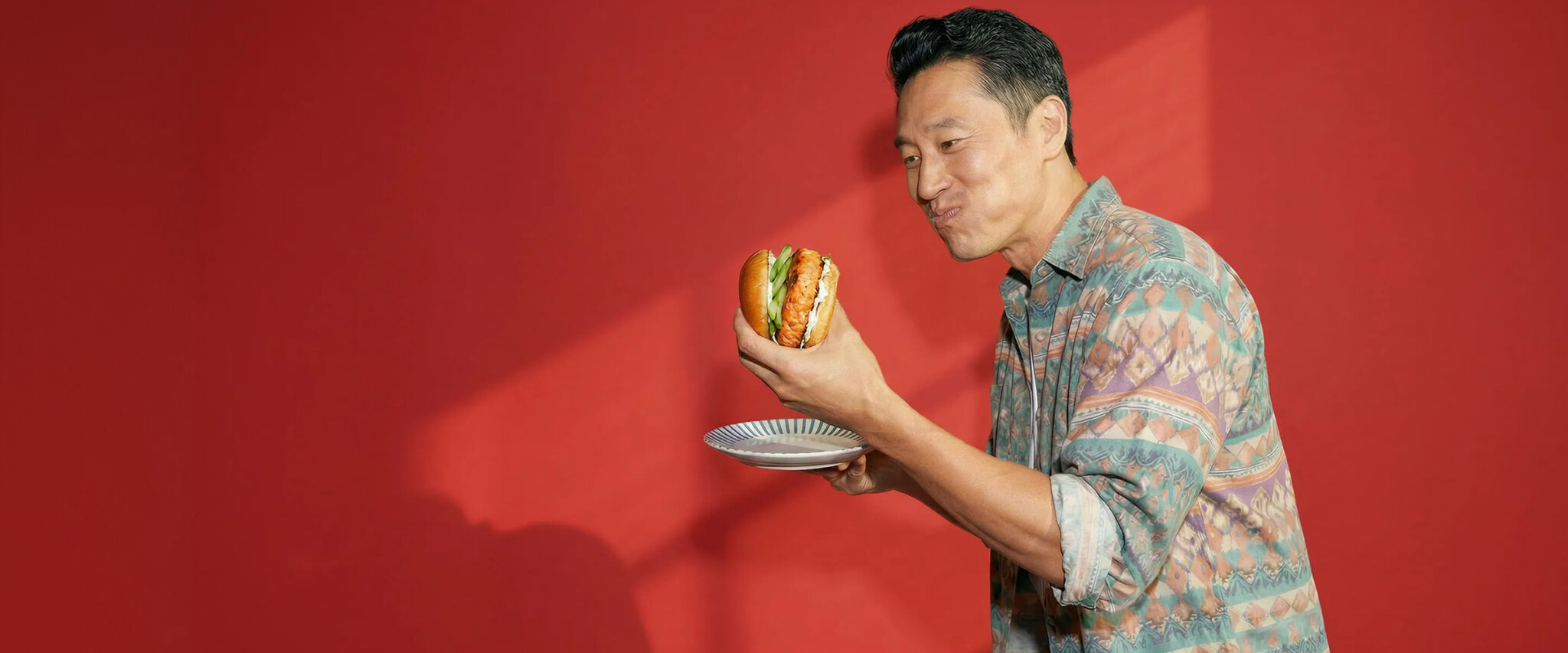 Uomo con camicia colorata che mangia un hamburger davanti a sfondo rosso.